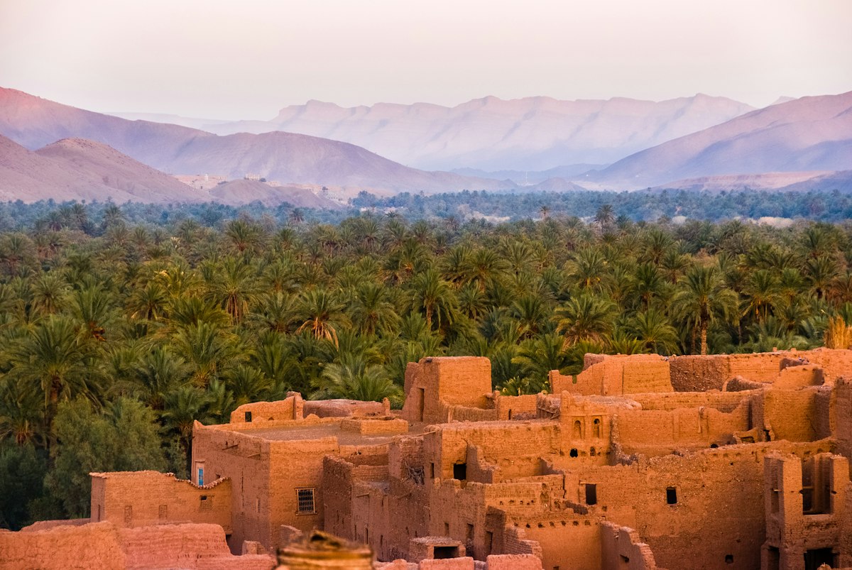 A Moroccan earthen kasbah of clay buildings overlooking a green palm oasis with the Atlas Mountains behind